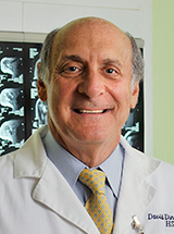 Person in a white coat and yellow tie smiles in front of medical imaging displays.