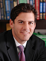 A man in a suit and tie smiles while sitting in front of a bookshelf.