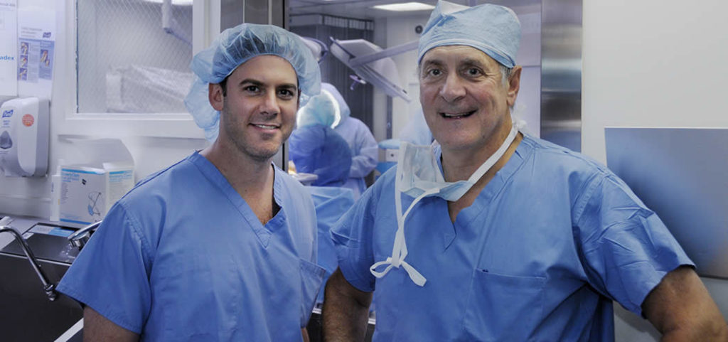 Two medical professionals in blue scrubs and surgical caps standing together in a hospital room.